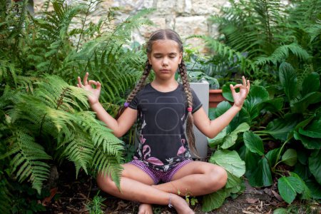 Foto de Una joven serena con el pelo trenzado medita en un jardín exuberante, rodeado de helechos vibrantes y plantas de hoja ancha. - Imagen libre de derechos