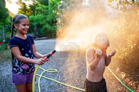 A girl smiles as she sprays her younger sibling with a garden hose, sparkling water drops glittering in the sunlight, encapsulating joyous summer fun