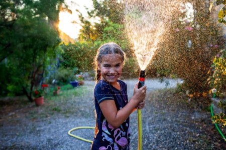 Foto de Una chica alegre juega con una manguera de jardín, rociando agua en la luz dorada del sol poniente, creando un rocío brumoso sobre un fondo de árboles. - Imagen libre de derechos