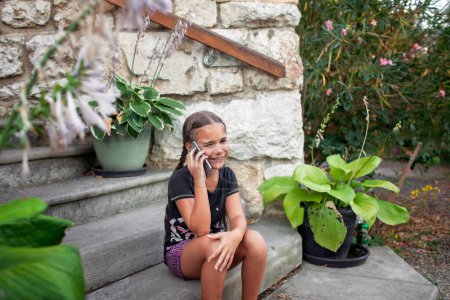 Foto de Una chica habla por teléfono mientras está sentada en escalones de piedra, rodeada de exuberante vegetación y plantas en maceta, encarnando la serenidad de unas vacaciones en una casa de campo. - Imagen libre de derechos