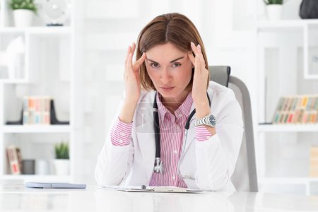 Téléchargez les photos : Portrait rapproché d'une femme médecin fatiguée assise sur le bureau et travaillant sur un ordinateur portable dans le bureau d'une clinique moderne - en image libre de droit