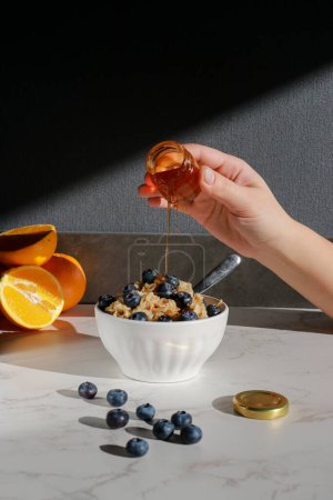Photo for Hand adding honey on oatmeal with blueberries on top in white porcelain bowl . White marble table and grey background - Royalty Free Image