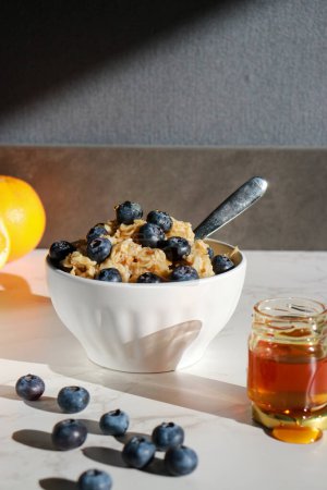 Photo for Healthy oatmeal with blueberries on top in white porcelain bowl, honey jar and oranges in background . White marble table and grey wall - Royalty Free Image