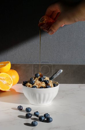 Photo for Hand adding honey on oatmeal with blueberries on top in white porcelain bowl . White marble table and grey background - Royalty Free Image