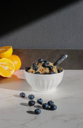 Photo for Healthy vegan oatmeal with blueberries in white porcelain bowl. White marble table and oranges in background - Royalty Free Image