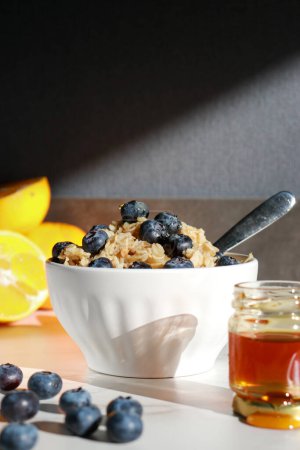 Photo for Healthy oatmeal with blueberries on top in white porcelain bowl, honey jar and oranges in background . White marble table and grey wall - Royalty Free Image