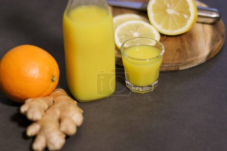 Draufsicht auf hausgemachtes Orangen-, Zitronen- und Ingwergetränk in Glasflasche mit kleinem Schnapsglas auf Küchentisch, verschwommener Hintergrund 