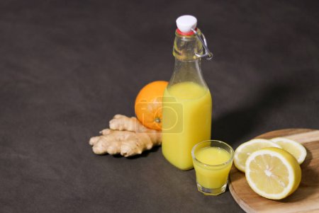 Draufsicht auf hausgemachtes Orangen-, Zitronen- und Ingwergetränk in Glasflasche mit kleinem Schnapsglas auf Küchentisch, verschwommener Hintergrund 