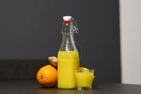 Hausgemachtes Orangen-, Zitronen- und Ingwergetränk in einer Glasflasche mit kleinem Schnapsglas auf dem Küchentisch, verschwommener Hintergrund 