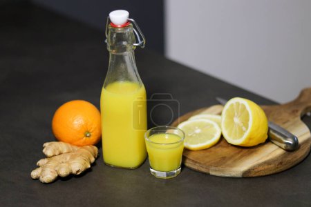 Homemade orange, lemon and ginger drink in a glass bottle with a small shot glass on kitchen table, blurry background 