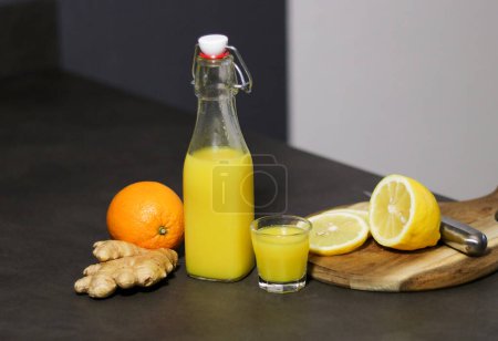Bebida casera de naranja, limón y jengibre en una botella de vidrio con un vaso pequeño en la mesa de la cocina, fondo borroso 