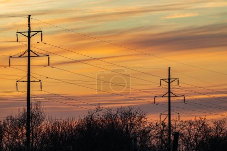 High-voltage overhead power line with wires and transmission tower (electricity pylon) poles against a sunset background