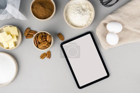 Tableta de pantalla táctil digital con ingredientes de receta brownie y utensilios de cocina sobre fondo gris, vista superior.