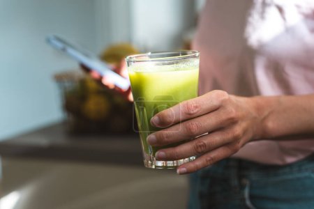 Photo for Unrecognizable woman drinks a fresh green healthy smoothie and uses a mobile phone. - Royalty Free Image