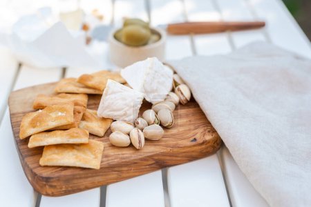 Foto de Aperitivos de vino a saber, pistachos, queso camembert, galletas saladas y aceitunas en una mesa blanca al aire libre. - Imagen libre de derechos