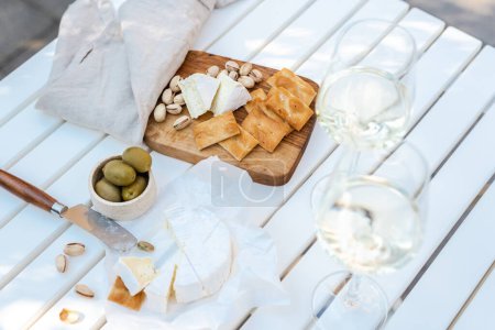 Foto de Dos copas de vino blanco y un plato de madera con queso y nueces en una mesa blanca al aire libre. - Imagen libre de derechos