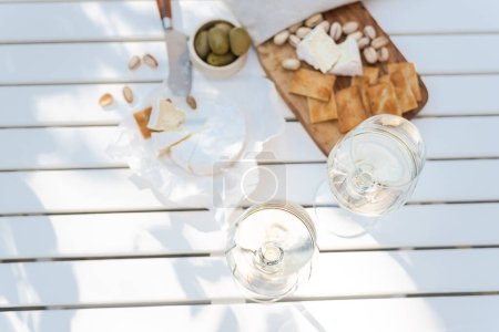 Photo for Two glasses of white wine and a wooden plate of cheese and nuts in the backyard. - Royalty Free Image