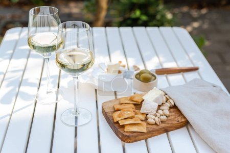 Two glasses of white wine and a wooden plate with cheese and nuts on a white table outdoors.