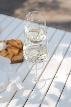 Photo for Two glasses of white wine and a wooden plate with cheese and nuts on a white table outdoors. - Royalty Free Image