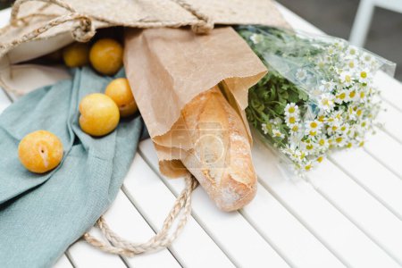 Bouquet of chamomile, ripe yellow plums, and a fresh baguette in a straw shopping bag.