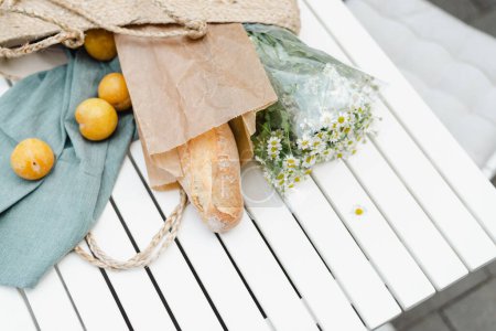 Foto de Ramo de manzanilla, ciruelas amarillas maduras y una baguette fresca en una bolsa de compras de paja. - Imagen libre de derechos