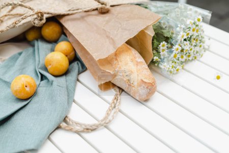 Foto de Ramo de manzanilla, ciruelas amarillas maduras y una baguette fresca en una bolsa de compras de paja. - Imagen libre de derechos