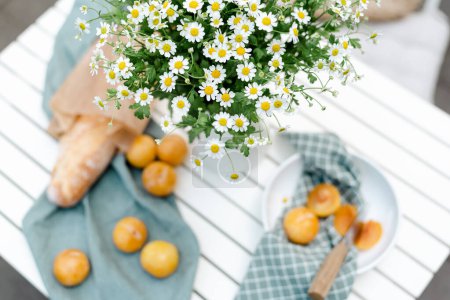 Foto de Ramo de manzanilla en una jarra blanca, ciruelas amarillas maduras y baguette fresca en una mesa blanca en el patio. - Imagen libre de derechos
