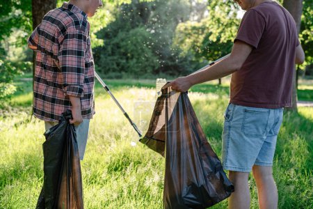 Téléchargez les photos : Les bénévoles ramassent une bouteille en plastique jetée de l'herbe dans un sac poubelle noir. - en image libre de droit