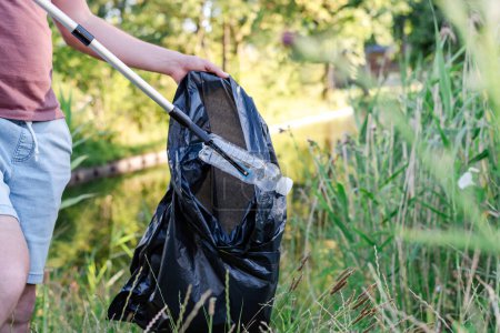 Téléchargez les photos : Un volontaire ramasse une bouteille en plastique jetée de l'herbe dans un sac poubelle noir. - en image libre de droit