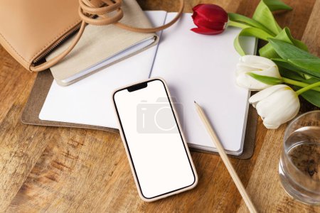 Foto de Vista superior del teléfono móvil con una pantalla blanca con lápiz, bloc de notas y bolso de moda en un escritorio. - Imagen libre de derechos