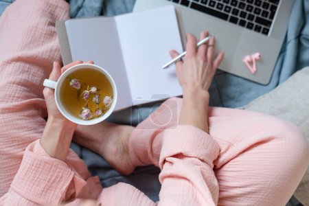 Téléchargez les photos : Une scène aux couleurs pastel d'une femme prenant des notes et buvant du thé chaud dans son lit. - en image libre de droit