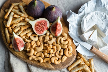 Wine appetizers namely ripe figs, camembert cheese, cashews, and salty bread sticks on the table indoors.
