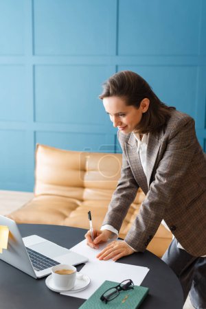 Belle femme brune debout au bureau, tenant un stylo et travaillant avec des documents.