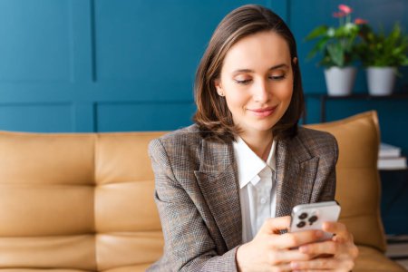 Photo for Beautiful woman with brown hair sitting on the sofa, smiling and reading a message on her mobile phone. - Royalty Free Image
