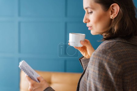 Schöne Geschäftsfrau steht im Büro, liest Notizen aus einem Notizblock und trinkt Kaffee.