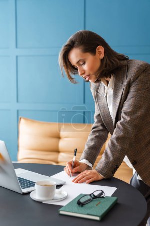 Schöne brünette Frau steht am Schreibtisch, hält Stift in der Hand und arbeitet mit Dokumenten.