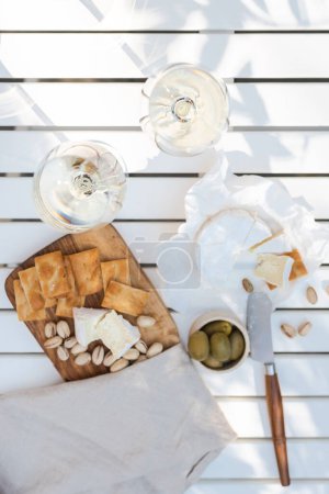Zwei Gläser Weißwein und ein Holzteller mit Käse und Nüssen auf einem weißen Tisch im Freien.