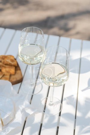 Deux verres de vin blanc et une assiette en bois avec fromage et noix sur une table blanche à l'extérieur.