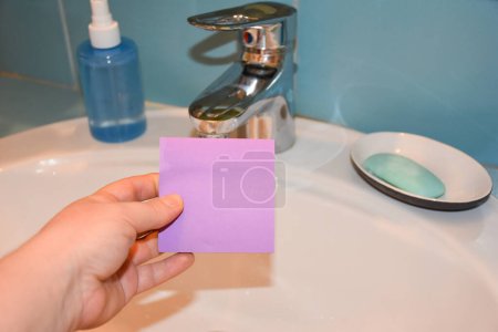 Photo for Woman hand holding a purple sticky note in front of a sink, bathroom. Covid-19 concept - Royalty Free Image