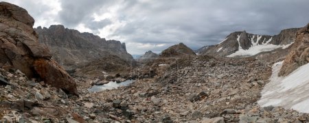 Photo for A panoramic shot of a talus field in the Alpine Lakes Basin in Wyoming's Wind River Range. The Brown Cliffs and the Fortress are seen in the distance. Three tiny hikers traverse the talus field. - Royalty Free Image