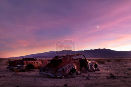 Téléchargez les photos : Le soleil se couche dans la vallée de Panamint dans le parc national de la vallée de la mort en Californie. De vieilles voitures rouillées et abandonnées sont visibles au premier plan. La chaîne de montagnes Argus est vue en arrière-plan. - en image libre de droit