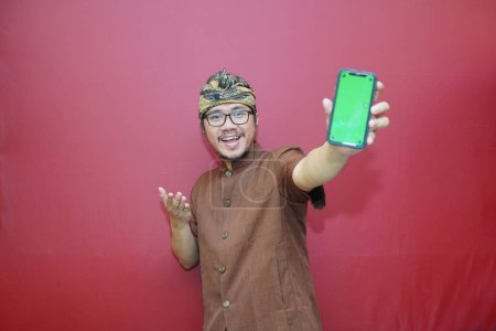 Foto de Excited Balinese man wearing udeng and and traditional clothes smiling showing screen of smartphone, isolated by red background. Gadget with blank empty space for mock up. - Imagen libre de derechos
