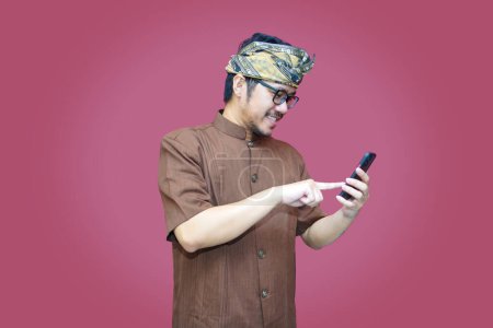 Foto de Un retrato de un hombre balinés feliz sonríe y sostiene su teléfono inteligente con udeng y ropa tradicional aislada por un fondo rojo. - Imagen libre de derechos