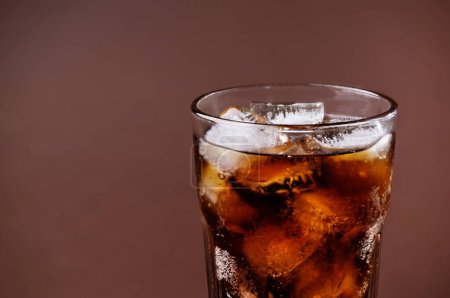 Téléchargez les photos : Un verre de boisson au cola avec des glaçons. Boisson fraîche de cola sucrée froide avec glace sur fond brun avec espace de copie. - en image libre de droit
