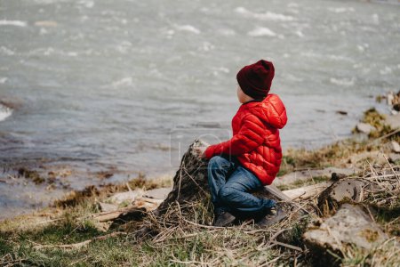 Foto de A little boy in a red jacket and a hat sits on a log by the river in spring. - Imagen libre de derechos