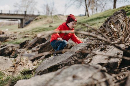 Foto de A little boy in a red jacket and hat climbs on a stone on the river bank in spring - Imagen libre de derechos
