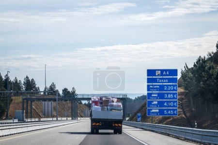 Blue information board for fees payable on the A4 motorway for different vehicle categories. Goods transport van passing on the highway.
