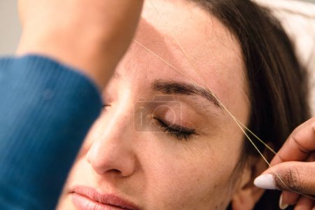 Caucasian woman removing hair from eyebrows with thread technique done by eyebrow stylist. Threading. Process Steps. Close up