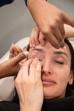 Kaukasische Frau entfernt die Haare von den Augenbrauen mit Fadentechnik, die von einem Augenbrauen-Stylisten ausgeführt wird. Einfädeln. Prozessschritte. Nahaufnahme
