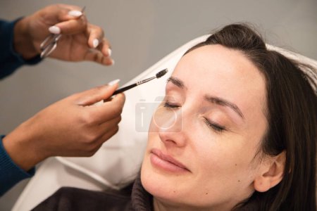 Kaukasische Frau entfernt die Haare von den Augenbrauen mit Fadentechnik, die von einem Augenbrauen-Stylisten ausgeführt wird. Einfädeln. Prozessschritte. Nahaufnahme